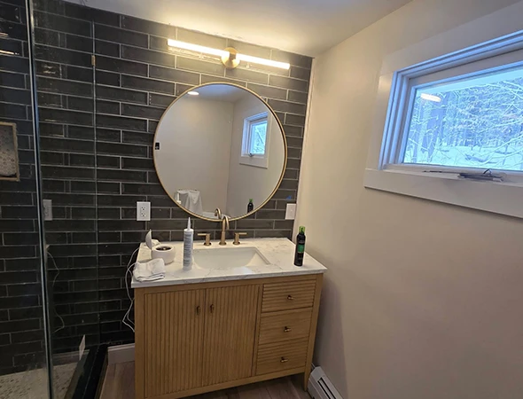 Full bathroom remodel in Burlington, VT, showcasing a walk-in glass shower with dark vertical tiles alongside a modern wood vanity and gold fixtures.