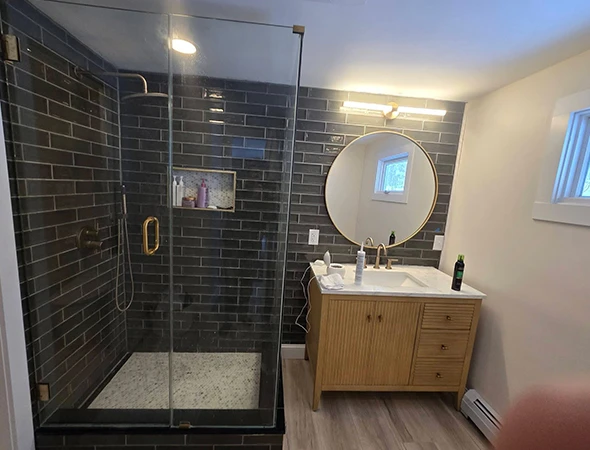 Modern bathroom vanity and sink upgrade in Newport, VT, featuring a light wood cabinet, gold-rimmed circular mirror, and dark tile accent wall.
