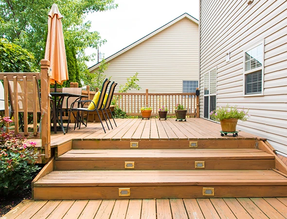 Custom composite deck building in Newport, VT, showing wide wooden stairs with integrated lighting, a patio dining set with an umbrella, and beige vinyl siding.